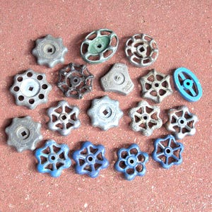 May include: A collection of seventeen metal and plastic valve handles in various shapes and sizes. The assortment includes round, star-shaped, and oval designs in shades of silver, grey, black, green, and blue, featuring central square holes.