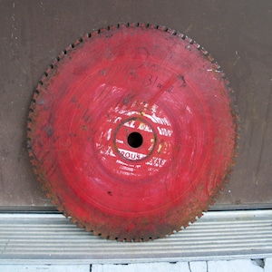 May include: A circular, red saw blade with a central hole and a toothed outer edge. The blade has some rust and wear, with the handwritten text "Dull Blade" visible. The center of the blade has printed text.