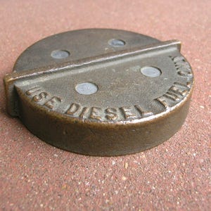 May include: A bronze-colored, circular fuel cap with the embossed text "USE DIESEL FUEL". The cap has a raised rectangular section and three small, evenly spaced holes. The cap is designed for diesel fuel.