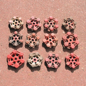 May include: A collection of twelve circular metal valve handles arranged in a grid. Each handle features a six-spoke design with a square center opening. The handles are finished in various shades of red, with some showing signs of wear.