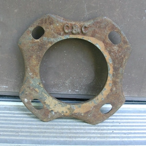 May include: A rusty, square metal flange with a central circular opening and four smaller holes. The letters "C&C" are embossed on the top. The metal has a weathered appearance with signs of corrosion and age.