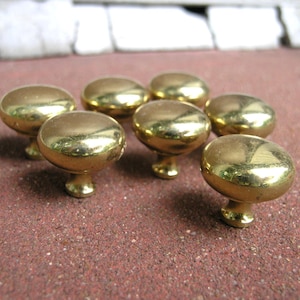 May include: Seven shiny, gold-colored cabinet knobs with a rounded top and a short, decorative base. The knobs are arranged on a reddish-brown surface, reflecting light. These knobs are suitable for furniture restoration or new builds.