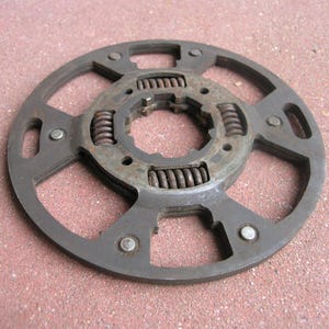 May include: A circular, dark gray metal component with a central hub and multiple springs. The component has a series of cut-out sections and is secured with rivets. The object appears to be a mechanical part, possibly for a machine or engine.