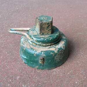 May include: A weathered, green metal object with a square top and a circular base. The object has a handle and shows signs of age with chipped paint and rust. The item appears to be a tool or part of a larger mechanism.