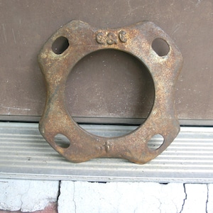May include: A rusty, circular metal flange with a central hole and four smaller holes around the perimeter. The flange has the letters "C&C" and the number "4" stamped on it. The metal has a weathered, aged appearance.