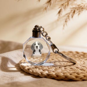 May include: A clear, faceted crystal keyring featuring a Beagle dog portrait. The dog has black and white markings and the name "Scout" is etched below. The keyring has a silver-coloured metal top and a keyring.