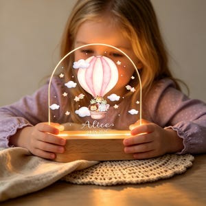 May include: A custom acrylic night light with a wooden base, featuring a printed illustration of a pink hot air balloon carrying a giraffe, lion, and elephant. The design includes clouds and stars, with the name Alice and date 24.05.2026 printed below.