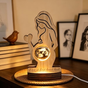 May include: A light-coloured wooden lamp shaped like a pregnant figure with long hair, featuring a glowing circular glass insert in the belly area and a heart-shaped cutout. The base is engraved with the text Love at first heart beat.