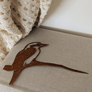 May include: A rusty brown metal sculpture of a kookaburra bird perched on a branch. The bird is detailed with cut-out features and sits on a light gray surface. The sculpture is a decorative item.