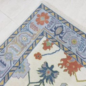 Puede incluir: Una esquina de una alfombra color crema con un dise&ntilde;o floral. El borde es azul claro con patrones geom&eacute;tricos amarillos y azul marino. La alfombra presenta motivos florales naranjas y azules.