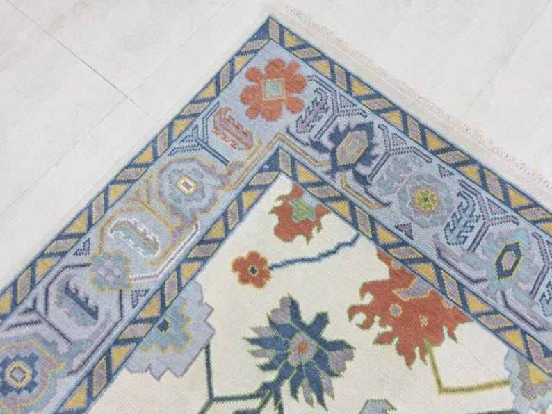 Puede incluir: Una esquina de una alfombra color crema con un dise&ntilde;o floral. El borde es azul claro con patrones geom&eacute;tricos amarillos y azul marino. La alfombra presenta motivos florales naranjas y azules.