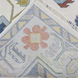 Puede incluir: Primer plano de una secci&oacute;n de una alfombra tejida con un dise&ntilde;o floral. La alfombra presenta un fondo crema con un borde en tonos azules, amarillos y melocot&oacute;n. El dise&ntilde;o incluye formas geom&eacute;tricas y un motivo floral central.