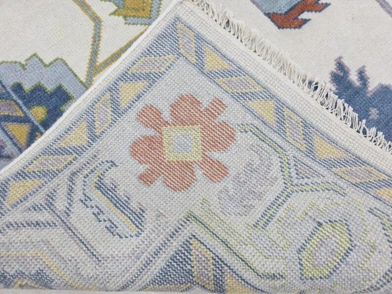 Puede incluir: Primer plano de una secci&oacute;n de una alfombra tejida con un dise&ntilde;o floral. La alfombra presenta un fondo crema con un borde en tonos azules, amarillos y melocot&oacute;n. El dise&ntilde;o incluye formas geom&eacute;tricas y un motivo floral central.