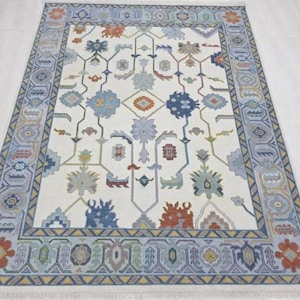 Puede incluir: Alfombra rectangular con un campo crema y un borde detallado. La alfombra presenta un patr&oacute;n central de motivos florales y geom&eacute;tricos estilizados en tonos azules, naranjas y verdes. El borde tiene un dise&ntilde;o geom&eacute;trico repetitivo.