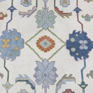Puede incluir: Primer plano de una alfombra crema con un patr&oacute;n repetitivo de formas florales y geom&eacute;tricas. El dise&ntilde;o presenta elementos azules, naranjas y verdes, creando una composici&oacute;n sim&eacute;trica y detallada. La textura de la alfombra es visible, sugiriendo una construcci&oacute;n tejida.