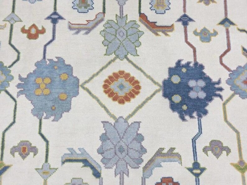 Puede incluir: Primer plano de una alfombra crema con un patr&oacute;n repetitivo de formas florales y geom&eacute;tricas. El dise&ntilde;o presenta elementos azules, naranjas y verdes, creando una composici&oacute;n sim&eacute;trica y detallada. La textura de la alfombra es visible, sugiriendo una construcci&oacute;n tejida.