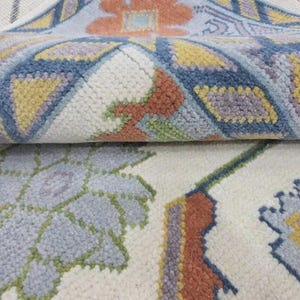 Puede incluir: Primer plano de una alfombra enrollada con un fondo crema y dise&ntilde;os florales y geom&eacute;tricos. El dise&ntilde;o incorpora azul, amarillo, naranja y verde. La textura de la alfombra parece suave y afelpada.