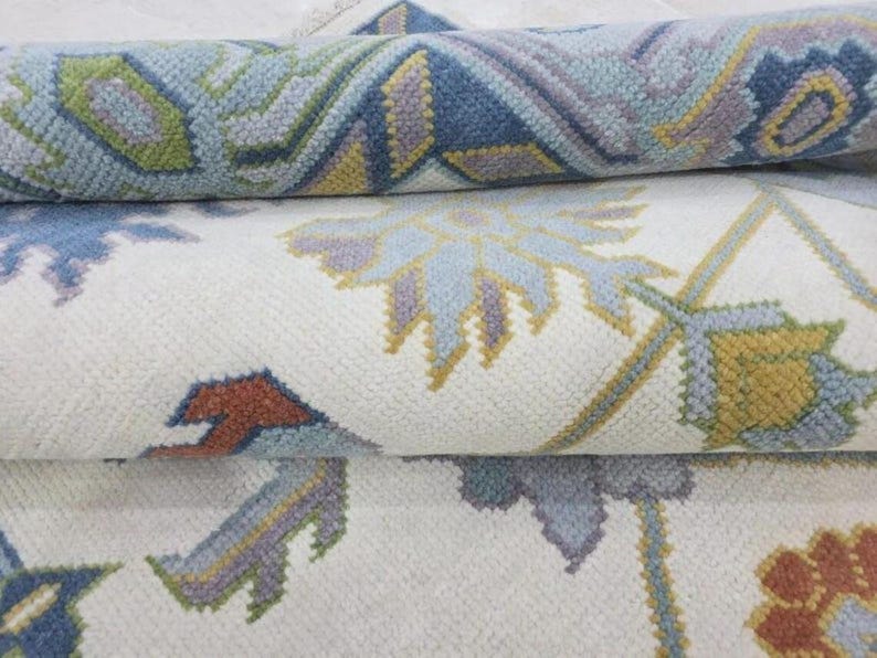 Puede incluir: Primer plano de una alfombra enrollada con un estampado floral. La alfombra presenta un fondo crema con dise&ntilde;os florales y geom&eacute;tricos azules, verdes, amarillos y &oacute;xido. La alfombra parece estar hecha de un material suave y tejido.