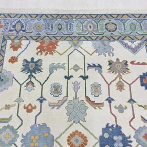 Puede incluir: Primer plano de una alfombra crema con un patr&oacute;n floral detallado. La alfombra presenta un borde en tonos azules, con acentos de naranja, rojo y verde. El dise&ntilde;o central incluye motivos florales estilizados en varios colores.