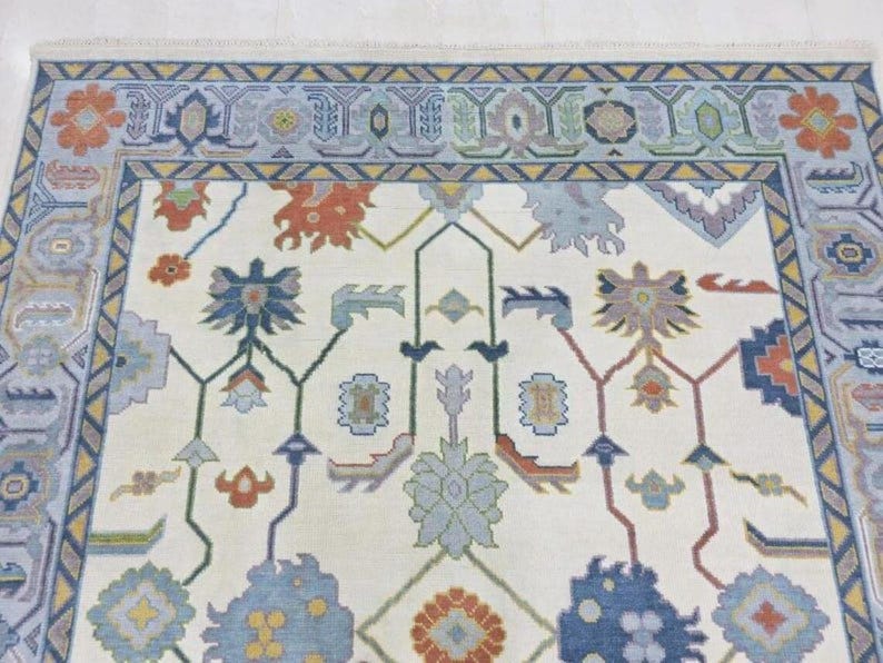 Puede incluir: Primer plano de una alfombra crema con un patr&oacute;n floral detallado. La alfombra presenta un borde en tonos azules, con acentos de naranja, rojo y verde. El dise&ntilde;o central incluye motivos florales estilizados en varios colores.