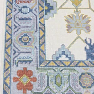 Puede incluir: Primer plano de una secci&oacute;n de una alfombra crema con un borde detallado. El borde presenta patrones geom&eacute;tricos en tonos azules, amarillos y naranjas. El dise&ntilde;o de la alfombra incluye motivos florales y geom&eacute;tricos.