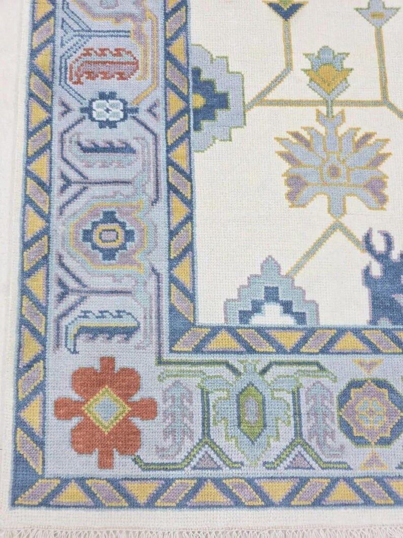 Puede incluir: Primer plano de una secci&oacute;n de una alfombra crema con un borde detallado. El borde presenta patrones geom&eacute;tricos en tonos azules, amarillos y naranjas. El dise&ntilde;o de la alfombra incluye motivos florales y geom&eacute;tricos.