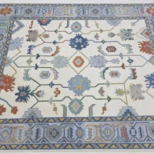 Puede incluir: Una alfombra rectangular con un campo color crema y un borde detallado. La alfombra presenta un patr&oacute;n floral repetitivo en tonos de azul, naranja y gris. El borde es un dise&ntilde;o geom&eacute;trico en azul y gris.