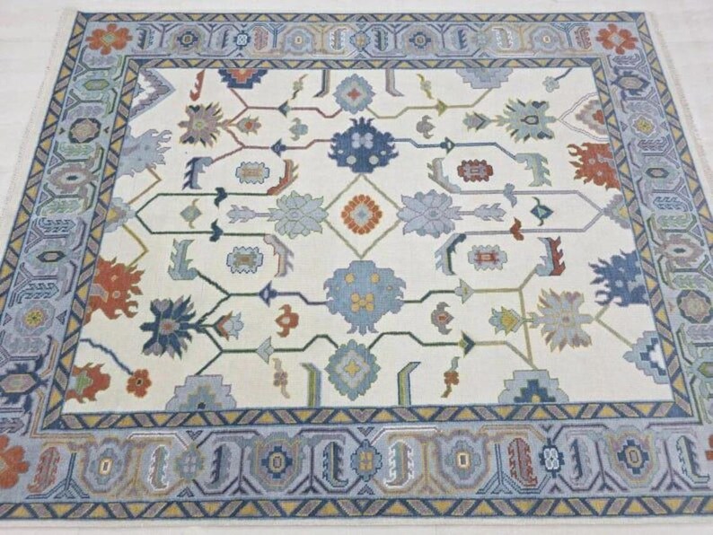 Puede incluir: Una alfombra rectangular con un campo color crema y un borde detallado. La alfombra presenta un patr&oacute;n floral repetitivo en tonos de azul, naranja y gris. El borde es un dise&ntilde;o geom&eacute;trico en azul y gris.