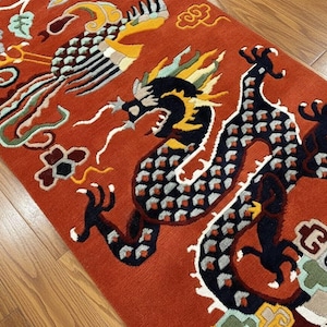 Op de afbeelding: Rood tapijt met een ingewikkeld ontwerp met een draak en een feniks. De draak is zwart, blauw en geel, terwijl de feniks gele, witte en zwarte details heeft. Het tapijt heeft een gefranjerde rand.