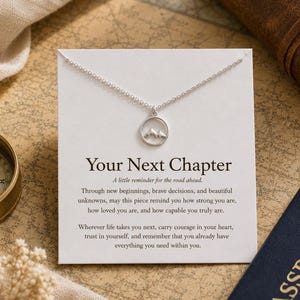 Puede incluir: Un collar de tono plateado con un colgante circular que presenta un diseño recortado de montañas. El collar se exhibe sobre una tarjeta blanca impresa con el texto Your Next Chapter y un mensaje inspirador sobre nuevos comienzos.