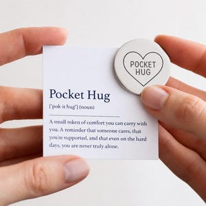 Może przedstawiać: Pocket Hug, mały symbol pocieszenia, trzymany w dłoniach. Na białej karcie napis "Pocket Hug" z definicją. Srebrny żeton w kształcie serca z napisem "Pocket Hug" znajduje się na górze karty.