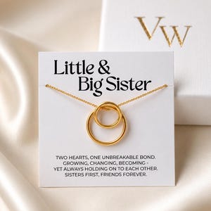 Pode incluir: Um colar dourado com dois círculos interligados, exibido em um cartão branco com a frase "Little & Big Sister". O cartão também inclui o texto: "Two hearts, one unbreakable bond."