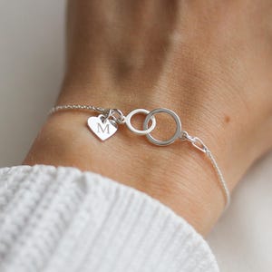 Peut inclure: Un bracelet en argent délicat avec deux cercles entrelacés et un charm en forme de cœur. Le charm en forme de cœur est gravé de la lettre "M". Le bracelet est porté au poignet, mettant en valeur sa fine chaîne et son design minimaliste.
