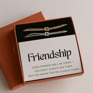Op de afbeelding: Een geschenkdoos met een set gouden en zilveren vriendschapsarmbanden. De armbanden hebben een knoopontwerp. De doos bevat een kaart met het woord "Vriendschap" en een citaat over vriendschap.