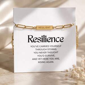 May include: A gold-colored chain bracelet with a rectangular charm that reads "RESILIENT." The bracelet is displayed on a white card with the word "Resilience" in large, bold letters. The card also includes inspirational text about overcoming challenges.