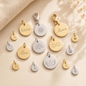 Könnte beinhalten: Eine Sammlung runder Anhänger aus gold- und silberfarbenem Metall mit verschiedenen Gravuren, darunter die Namen Emma und Mama, die Buchstaben A, M, K, R, ein Gänseblümchen, ein Regenbogen, ein Unendlichkeitssymbol, ein Herz, eine Pfote, Flügel und ein Kreuz.