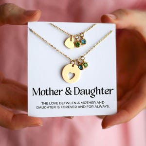 Peut inclure: Deux colliers dorés sur une carte blanche. L'un a un pendentif en forme de cœur avec une découpe en forme de cœur, l'autre a un pendentif en forme de cœur. Les deux colliers ont des breloques en pierres précieuses. La carte indique "Mother & Daughter".