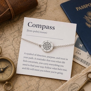 May include: A silver compass charm bracelet with a round pendant featuring a compass rose design. The bracelet is displayed on a card with the word "Compass" and a definition. A passport and ticket are in the background.