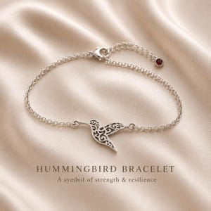 Könnte beinhalten: Silbernes Armband mit einem filigranen Kolibri-Anhänger und einem kleinen roten Edelstein. Das Armband hat eine feine Kette. Der Text auf dem Bild lautet "HUMMINGBIRD BRACELET" und "A symbol of strength & resilience."