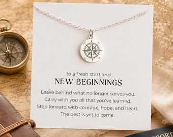 May include: A silver compass pendant necklace on a white card with the words "to a fresh start and NEW BEGINNINGS". The card is on a map with a compass and passport in the background. The necklace is a round pendant with a compass rose design.