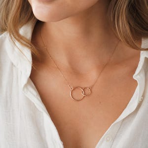 May include: A rose gold necklace with two interlinked circles. The fine chain and pendant are set against a white shirt. The circles are joined, representing unity. This necklace is a simple, elegant piece of jewellery, suitable for everyday wear.