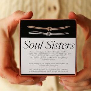 Może przedstawiać: Pudełko upominkowe z napisem "Soul Sisters" i dwiema pasującymi bransoletkami z węzłem, jedną złotą i jedną srebrną. Pudełko zawiera również wiersz o więzi przyjaźni.