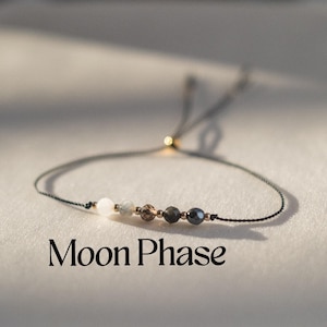 May include: A delicate bracelet featuring a series of small, multi-colored beads strung on a thin, dark cord. The beads include white, gray, brown, and dark gray stones. The text "Moon Phase" is displayed below the bracelet.