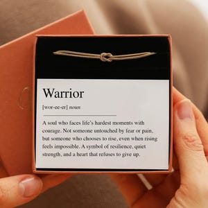 Puede incluir: Una pulsera de cadena dorada con un diseño de nudo central, presentada en una caja con forro negro y una tarjeta blanca. La tarjeta muestra el texto: Warrior [wor-ee-er] noun. Un alma que enfrenta los momentos más difíciles con valentía.