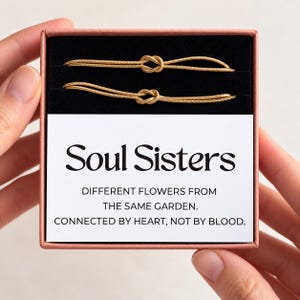 Op de afbeelding: Twee goudkleurige slangenketting-armbanden met centrale knoopdetails, gepresenteerd in een vierkante doos met een zwarte fluwelen inzet. Een witte kaart bevat de tekst: Soul Sisters, DIFFERENT FLOWERS FROM THE SAME GARDEN. CONNECTED BY HEART, NOT BY BLOOD.