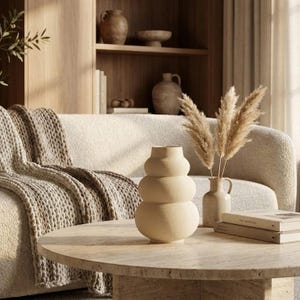 May include: A neutral-toned living room scene featuring a round coffee table with a stacked vase and a small vase with dried pampas grass. A beige sofa with a knitted throw and a wooden bookshelf in the background complete the decor.