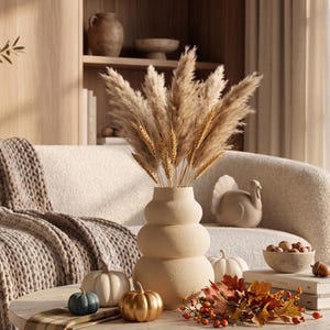 Puede incluir: Un jarrón de cerámica beige de varios niveles redondeados, lleno de hierba de la pampa alta y plumosa y tallos de trigo dorados. El jarrón se encuentra entre elementos decorativos que incluyen pequeñas calabazas en blanco, azul y dorado, hojas de otoño y una figura de pavo.