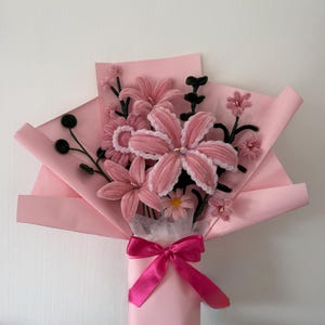 Puede incluir: Un ramo de flores rosas de hilo, envuelto en papel rosa y atado con una cinta de raso rosa. El ramo presenta lirios y margaritas, con detalles verdes. Un arreglo floral encantador.