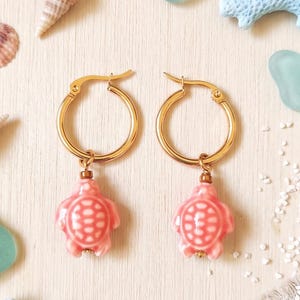 Può includere: Un paio di orecchini a cerchio color oro con piccoli ciondoli a forma di tartaruga rosa. Ogni ciondolo presenta un motivo a guscio dettagliato ed è sospeso a un cerchio di metallo circolare con un meccanismo di chiusura a cerniera.