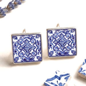Könnte beinhalten: Ein Paar quadratische Ohrstecker mit einem komplizierten blau-weißen Blumenmuster in einer silberfarbenen Metallfassung. Zwei kleine quadratische Keramikfliesen mit passendem blau-weißem Design liegen daneben, zusammen mit Lavendelzweigen.